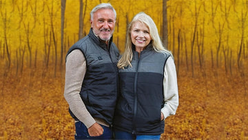 A man and woman wearing heated gilets, standing together in front of an autumn forest