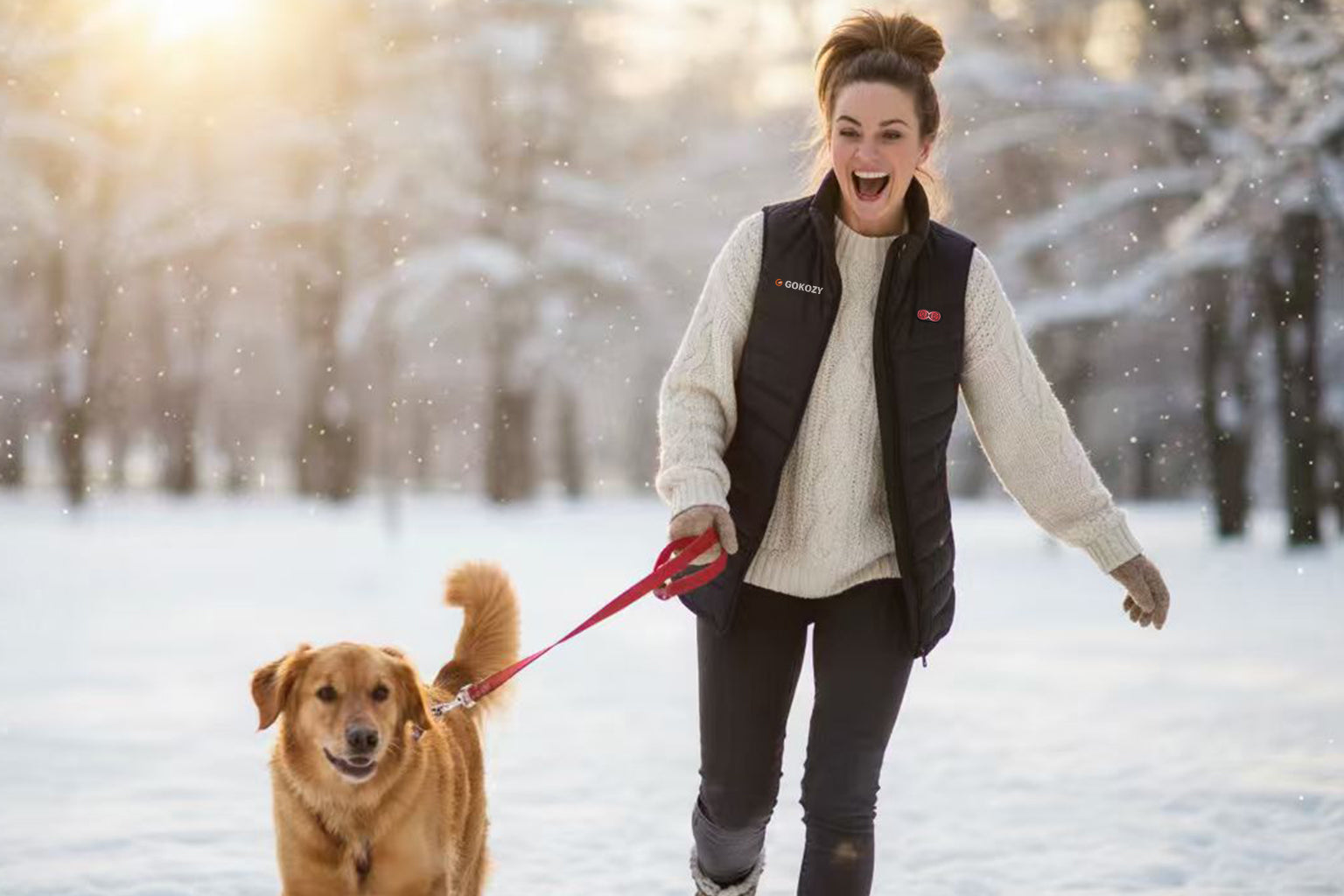  A Caucasian woman in her 30s wearing a black heated vest smiles while being gently pulled by her dog on a snowy neighborhood walk, her eyes are open and cheerful.